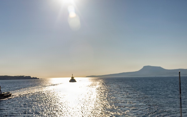 A large naval or military vessel is navigating coastal waters, viewed from its deck, with a tugboat escorting it on the port side. The scene is backlit by a low sun creating a bright glare path across the water, with mountainous coastline visible in the background. Several crew members are visible on the deck, suggesting a port departure or arrival operation.