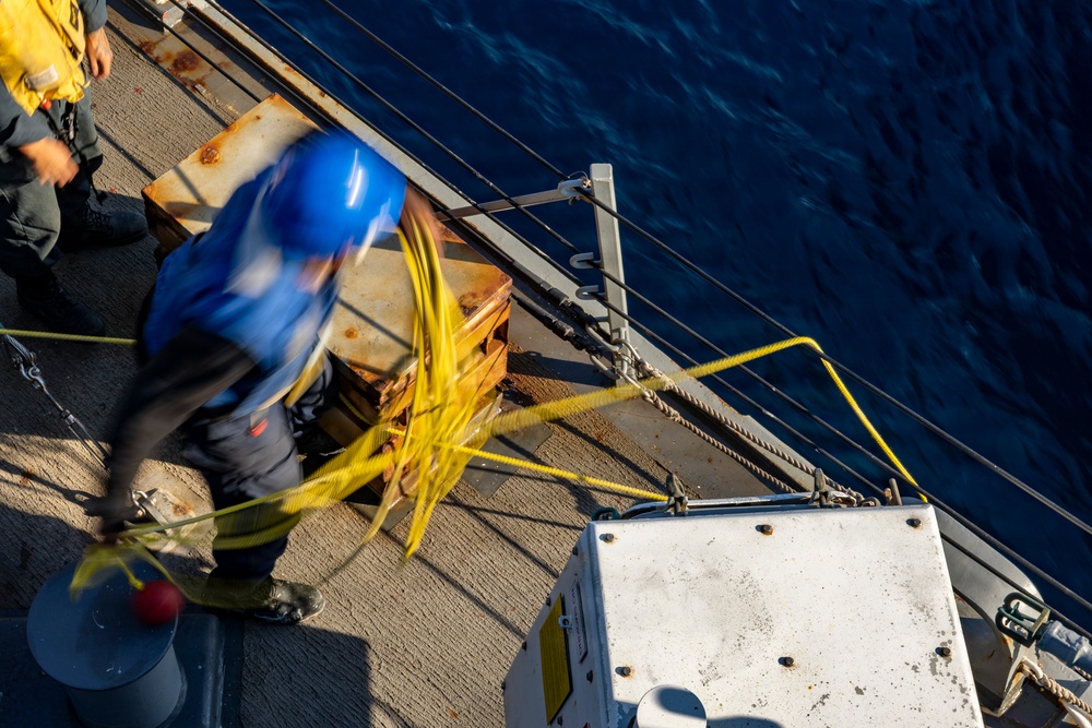 An aerial view of a ship's deck showing a worker in a blue hard hat handling yellow rope or cable near the vessel's edge. The image is taken from above, revealing the dark blue ocean water alongside the ship. A second person in a yellow jacket is partially visible in the upper left corner.