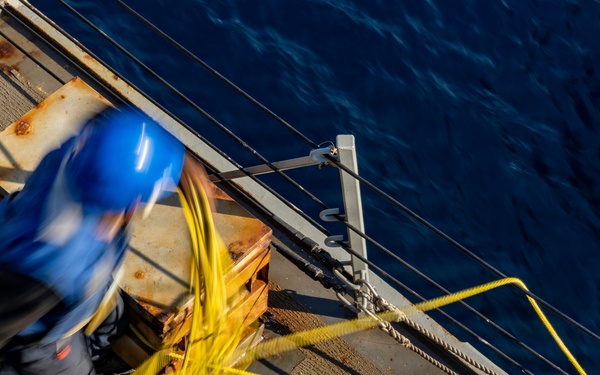 An aerial view of a ship's deck showing a worker in a blue hard hat handling yellow rope or cable near the vessel's edge. The image is taken from above, revealing the dark blue ocean water alongside the ship. A second person in a yellow jacket is partially visible in the upper left corner.