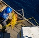 An aerial view of a ship's deck showing a worker in a blue hard hat handling yellow rope or cable near the vessel's edge. The image is taken from above, revealing the dark blue ocean water alongside the ship. A second person in a yellow jacket is partially visible in the upper left corner.