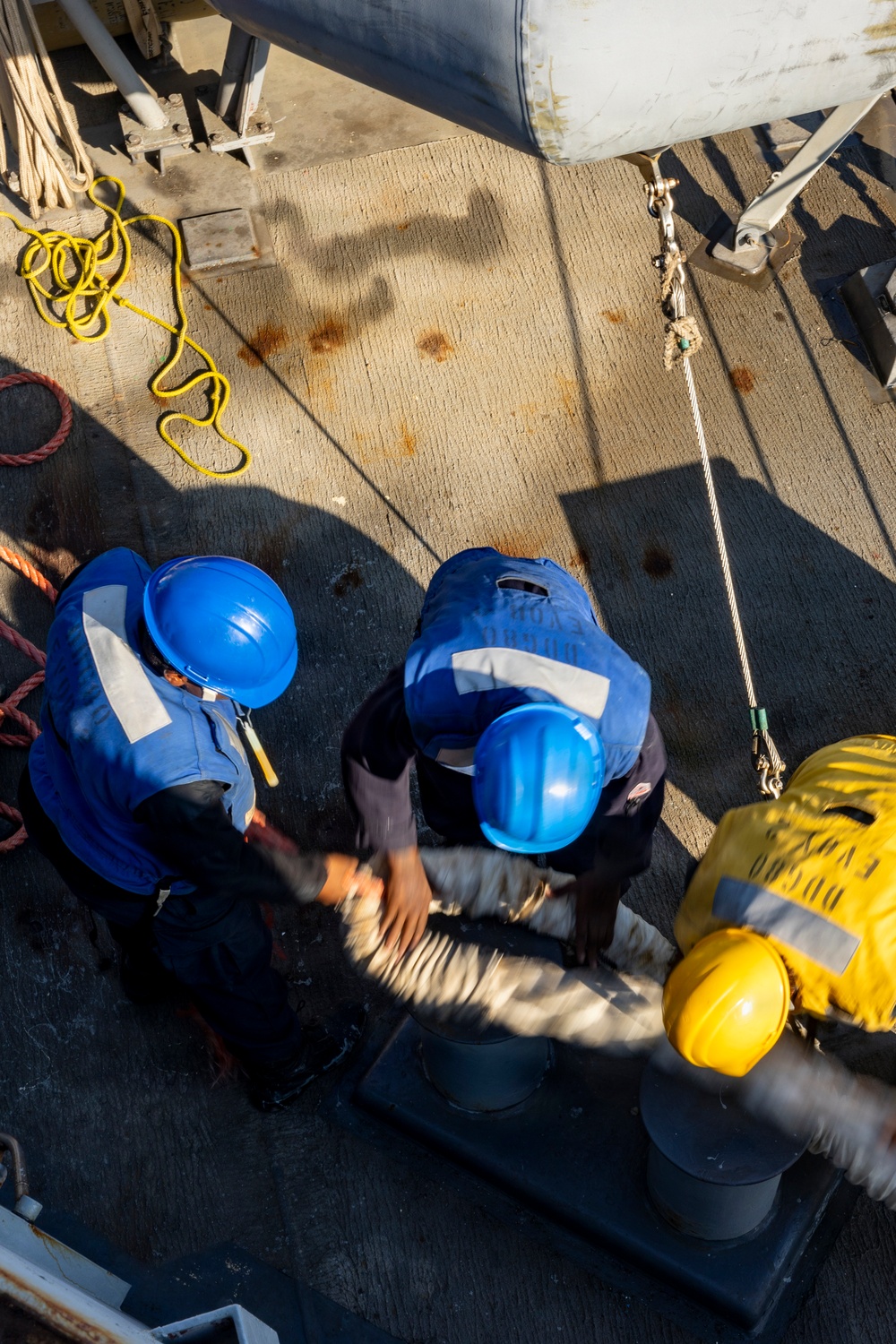 An overhead/bird's-eye view of workers on what appears to be a ship or industrial vessel deck. Two workers wearing blue hard hats and blue high-visibility vests are bent over handling equipment, while a third worker in a yellow hard hat and yellow gear is partially visible at the right edge. The deck shows signs of wear with rust stains, coiled ropes, and various rigging hardware.