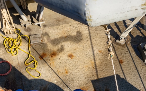 An overhead/bird's-eye view of workers on what appears to be a ship or industrial vessel deck. Two workers wearing blue hard hats and blue high-visibility vests are bent over handling equipment, while a third worker in a yellow hard hat and yellow gear is partially visible at the right edge. The deck shows signs of wear with rust stains, coiled ropes, and various rigging hardware.