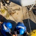An overhead/bird's-eye view of workers on what appears to be a ship or industrial vessel deck. Two workers wearing blue hard hats and blue high-visibility vests are bent over handling equipment, while a third worker in a yellow hard hat and yellow gear is partially visible at the right edge. The deck shows signs of wear with rust stains, coiled ropes, and various rigging hardware.