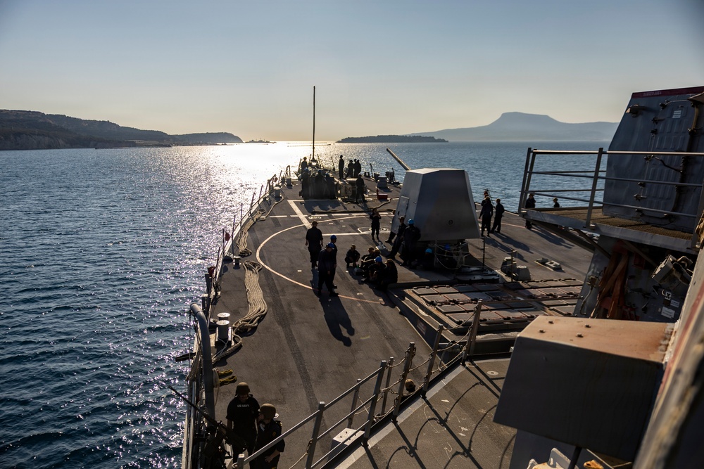 A U.S. Navy warship, identifiable by 'NAVY' markings on crew uniforms, is underway on calm blue waters near a coastline with hills and a flat-topped mountain visible in the background. The image is taken from an elevated position on the ship looking forward along the main deck toward the bow. The scene appears to be captured during early morning or late afternoon based on the low-angle sunlight reflecting off the water.