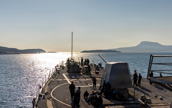 A U.S. Navy warship, identifiable by 'NAVY' markings on crew uniforms, is underway on calm blue waters near a coastline with hills and a flat-topped mountain visible in the background. The image is taken from an elevated position on the ship looking forward along the main deck toward the bow. The scene appears to be captured during early morning or late afternoon based on the low-angle sunlight reflecting off the water.