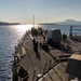 A U.S. Navy warship, identifiable by 'NAVY' markings on crew uniforms, is underway on calm blue waters near a coastline with hills and a flat-topped mountain visible in the background. The image is taken from an elevated position on the ship looking forward along the main deck toward the bow. The scene appears to be captured during early morning or late afternoon based on the low-angle sunlight reflecting off the water.