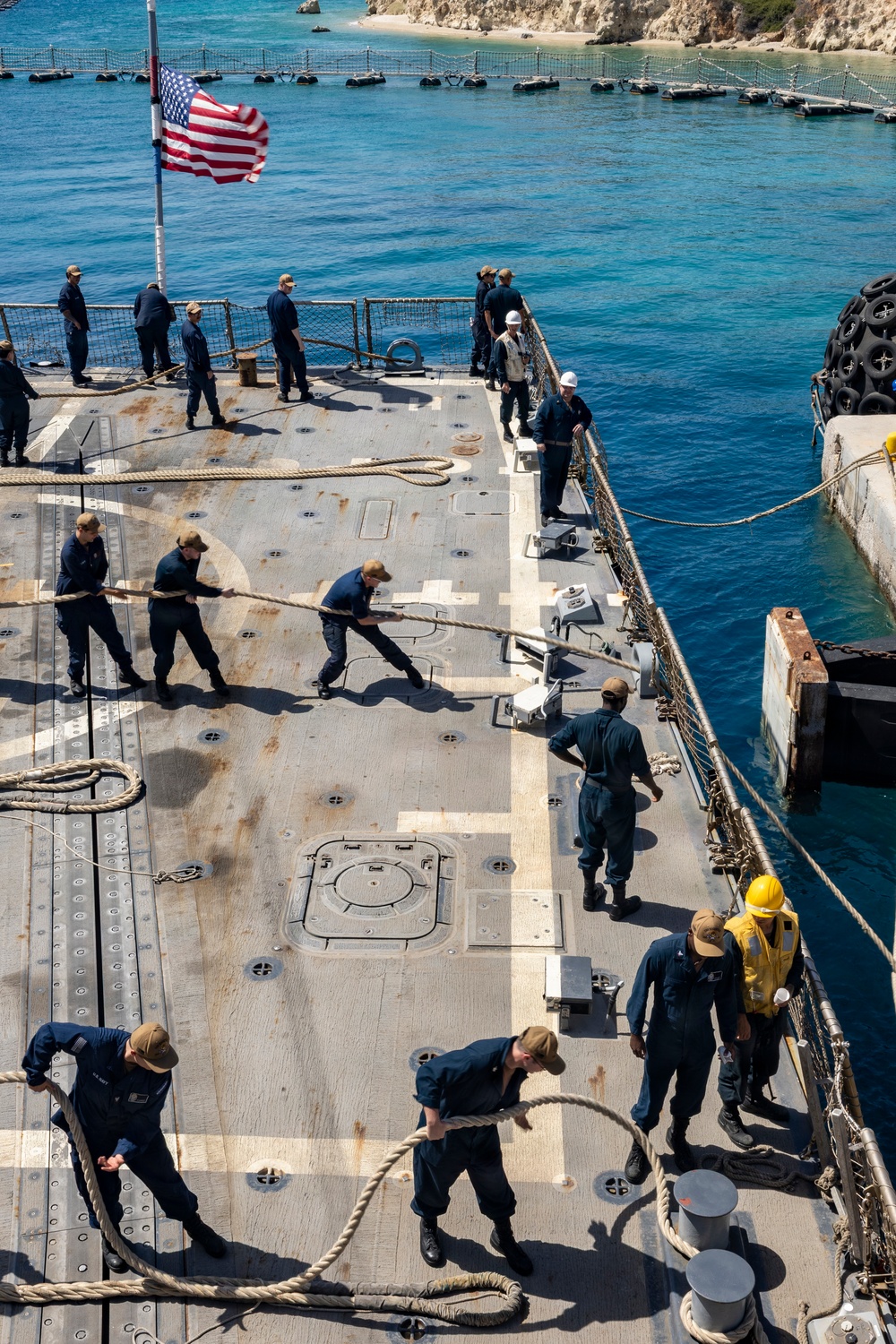 U.S. Navy sailors are conducting a mooring or line-handling evolution on the deck of a naval vessel, likely a destroyer or amphibious ship, as it comes alongside a pier or causeway. An American flag is flying from a mast, and the ship appears to be operating in clear blue coastal waters near a rocky shoreline. Several personnel in hard hats and safety vests are also present, suggesting a coordinated docking operation.