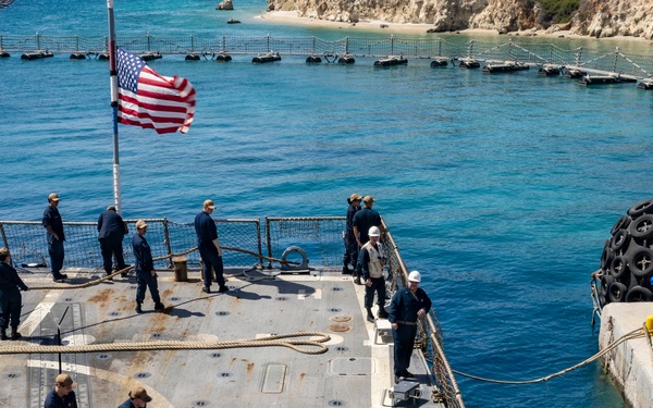 U.S. Navy sailors are conducting a mooring or line-handling evolution on the deck of a naval vessel, likely a destroyer or amphibious ship, as it comes alongside a pier or causeway. An American flag is flying from a mast, and the ship appears to be operating in clear blue coastal waters near a rocky shoreline. Several personnel in hard hats and safety vests are also present, suggesting a coordinated docking operation.