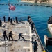 U.S. Navy sailors are conducting a mooring or line-handling evolution on the deck of a naval vessel, likely a destroyer or amphibious ship, as it comes alongside a pier or causeway. An American flag is flying from a mast, and the ship appears to be operating in clear blue coastal waters near a rocky shoreline. Several personnel in hard hats and safety vests are also present, suggesting a coordinated docking operation.