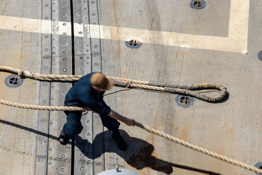 An aerial/overhead view of a person on what appears to be a ship's wooden deck handling large mooring or docking ropes. The individual is dressed in dark work clothing and is actively managing a thick braided rope with a looped end. The deck features metal track rails, bolted fittings, deck cleats, and painted markings consistent with a naval or commercial vessel.