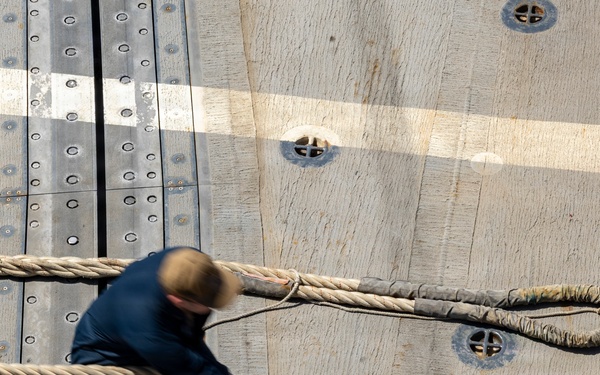 An aerial/overhead view of a person on what appears to be a ship's wooden deck handling large mooring or docking ropes. The individual is dressed in dark work clothing and is actively managing a thick braided rope with a looped end. The deck features metal track rails, bolted fittings, deck cleats, and painted markings consistent with a naval or commercial vessel.
