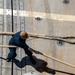 An aerial/overhead view of a person on what appears to be a ship's wooden deck handling large mooring or docking ropes. The individual is dressed in dark work clothing and is actively managing a thick braided rope with a looped end. The deck features metal track rails, bolted fittings, deck cleats, and painted markings consistent with a naval or commercial vessel.