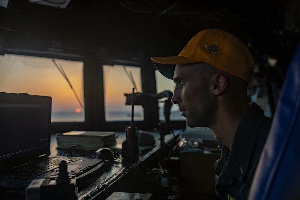 A young male crew member is seated at the bridge/helm of a naval or maritime vessel, focused on navigation equipment during what appears to be a sunset or sunrise. He is wearing a yellow baseball cap with a compass rose or star emblem and a dark uniform. The scene captures a working maritime environment with multiple electronic and communication devices visible.