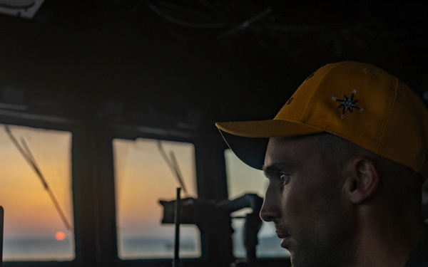 A young male crew member is seated at the bridge/helm of a naval or maritime vessel, focused on navigation equipment during what appears to be a sunset or sunrise. He is wearing a yellow baseball cap with a compass rose or star emblem and a dark uniform. The scene captures a working maritime environment with multiple electronic and communication devices visible.
