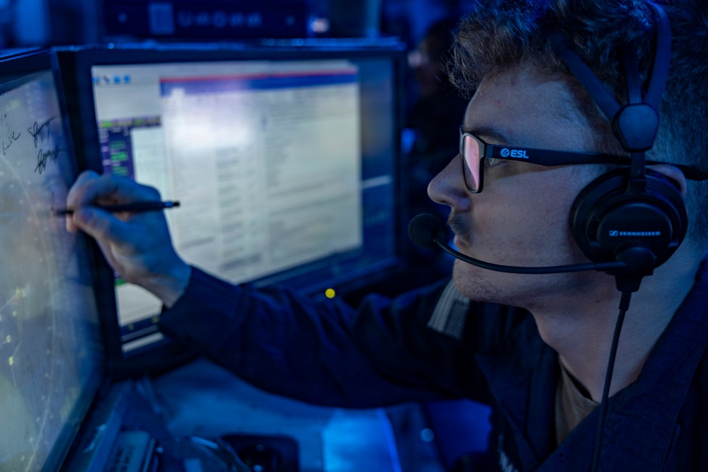 A young man wearing ESL-branded Sennheiser headphones and glasses is seated at a multi-monitor workstation in a dimly blue-lit environment. He is writing or drawing on a touchscreen monitor with a stylus while appearing to communicate via the headset microphone. The setting suggests an esports production, commentary, or analyst booth environment.