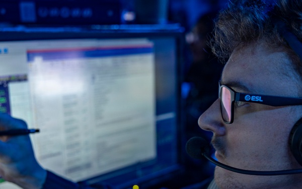 A young man wearing ESL-branded Sennheiser headphones and glasses is seated at a multi-monitor workstation in a dimly blue-lit environment. He is writing or drawing on a touchscreen monitor with a stylus while appearing to communicate via the headset microphone. The setting suggests an esports production, commentary, or analyst booth environment.