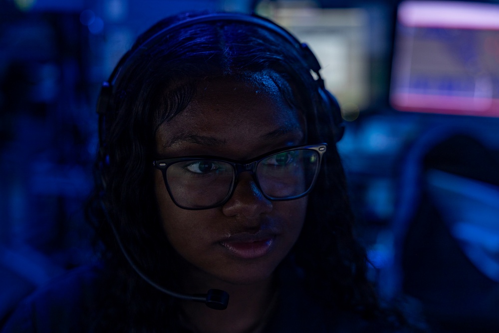 A young woman wearing a headset with a boom microphone and rectangular-framed glasses is photographed in a dimly lit environment with blue ambient lighting. She appears to be actively speaking or communicating, likely in a professional or gaming/esports context. Multiple illuminated monitors are visible in the blurred background.