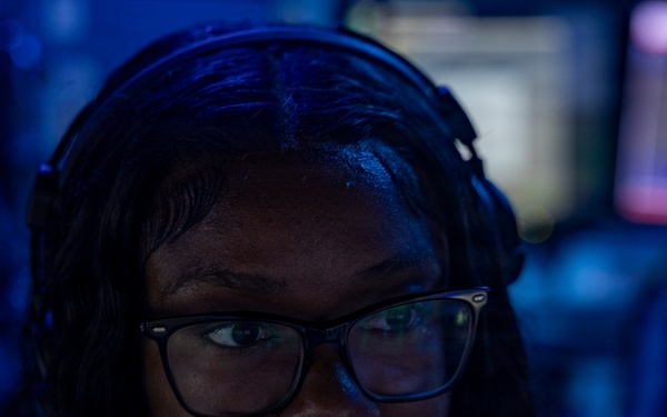 A young woman wearing a headset with a boom microphone and rectangular-framed glasses is photographed in a dimly lit environment with blue ambient lighting. She appears to be actively speaking or communicating, likely in a professional or gaming/esports context. Multiple illuminated monitors are visible in the blurred background.