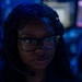 A young woman wearing a headset with a boom microphone and rectangular-framed glasses is photographed in a dimly lit environment with blue ambient lighting. She appears to be actively speaking or communicating, likely in a professional or gaming/esports context. Multiple illuminated monitors are visible in the blurred background.