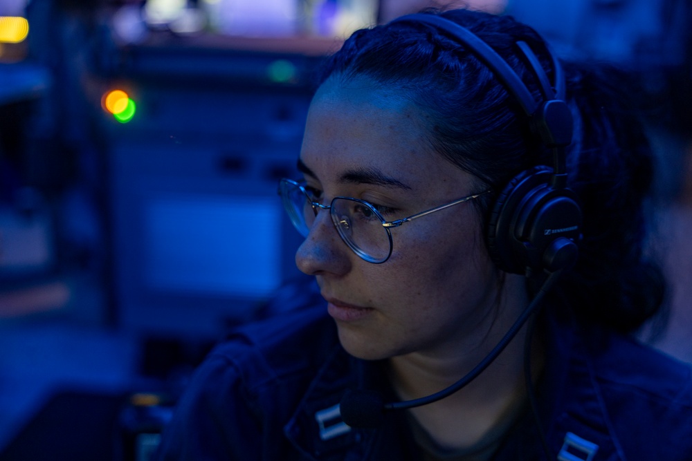 A person wearing military-style dark clothing, round-frame glasses, and black Sennheiser headphones is focused on a task in a dimly lit, blue-toned environment consistent with a naval operations center or combat information center. The individual has their hair pulled back and appears to be monitoring equipment or communications. The low-light blue ambient environment and visible indicator lights suggest an active duty watch station aboard a naval vessel.