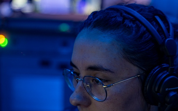 A person wearing military-style dark clothing, round-frame glasses, and black Sennheiser headphones is focused on a task in a dimly lit, blue-toned environment consistent with a naval operations center or combat information center. The individual has their hair pulled back and appears to be monitoring equipment or communications. The low-light blue ambient environment and visible indicator lights suggest an active duty watch station aboard a naval vessel.