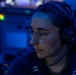 A person wearing military-style dark clothing, round-frame glasses, and black Sennheiser headphones is focused on a task in a dimly lit, blue-toned environment consistent with a naval operations center or combat information center. The individual has their hair pulled back and appears to be monitoring equipment or communications. The low-light blue ambient environment and visible indicator lights suggest an active duty watch station aboard a naval vessel.