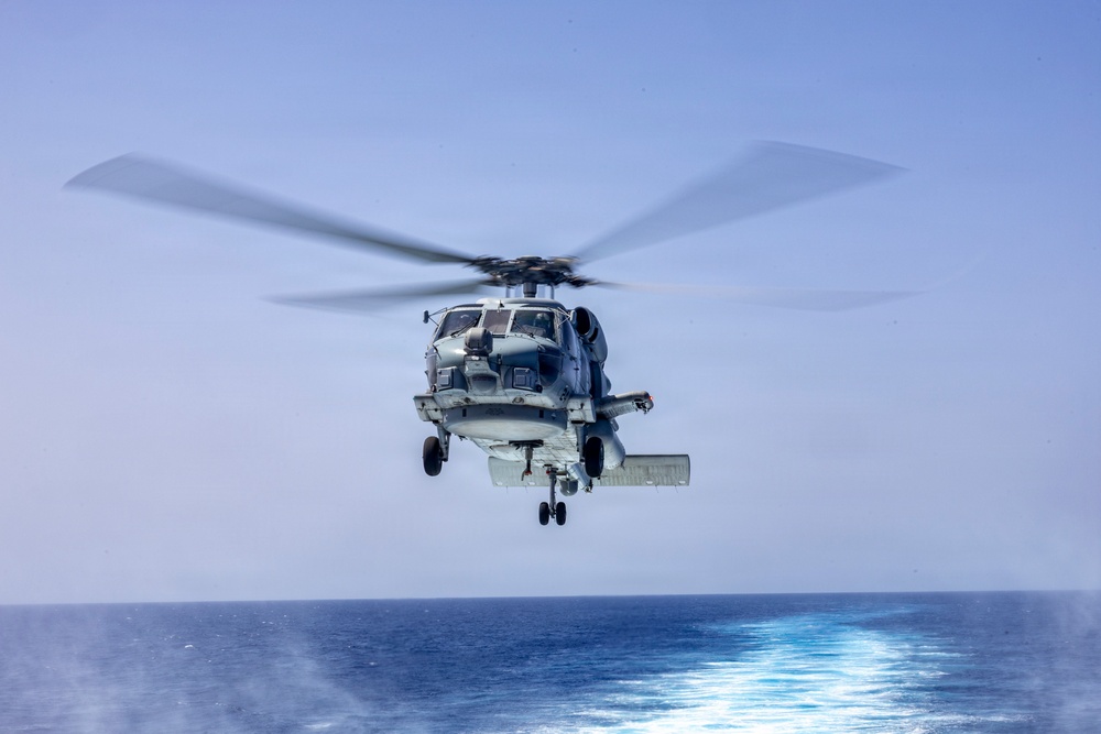 A military naval helicopter is captured in flight over open ocean, photographed from what appears to be the deck of a ship. The helicopter is shown in a low hover or approach position with its main rotor blades in motion, creating a motion blur effect. The setting is a clear day over calm, deep blue ocean waters.
