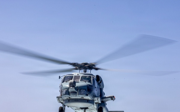 A military naval helicopter is captured in flight over open ocean, photographed from what appears to be the deck of a ship. The helicopter is shown in a low hover or approach position with its main rotor blades in motion, creating a motion blur effect. The setting is a clear day over calm, deep blue ocean waters.