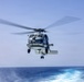 A military naval helicopter is captured in flight over open ocean, photographed from what appears to be the deck of a ship. The helicopter is shown in a low hover or approach position with its main rotor blades in motion, creating a motion blur effect. The setting is a clear day over calm, deep blue ocean waters.