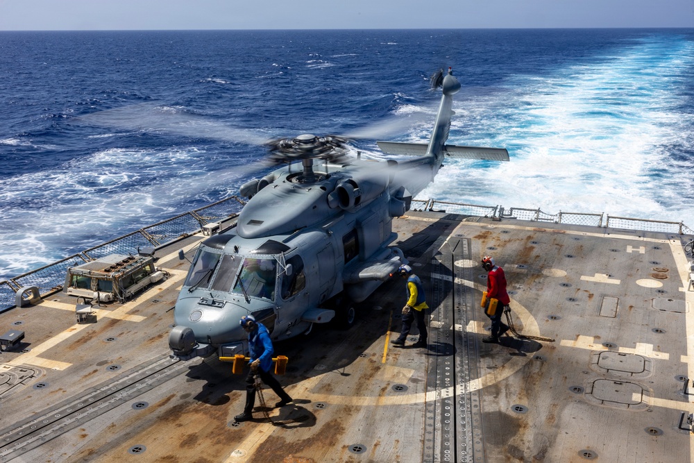 A military helicopter, consistent with an MH-60 Seahawk, is on the flight deck of a naval warship at sea with rotors spinning. Ground crew members in color-coded jerseys are performing deck operations around the aircraft. The ship is underway, evidenced by the visible wake trailing behind in open blue ocean water.