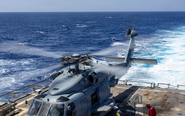 A military helicopter, consistent with an MH-60 Seahawk, is on the flight deck of a naval warship at sea with rotors spinning. Ground crew members in color-coded jerseys are performing deck operations around the aircraft. The ship is underway, evidenced by the visible wake trailing behind in open blue ocean water.