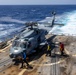 A military helicopter, consistent with an MH-60 Seahawk, is on the flight deck of a naval warship at sea with rotors spinning. Ground crew members in color-coded jerseys are performing deck operations around the aircraft. The ship is underway, evidenced by the visible wake trailing behind in open blue ocean water.