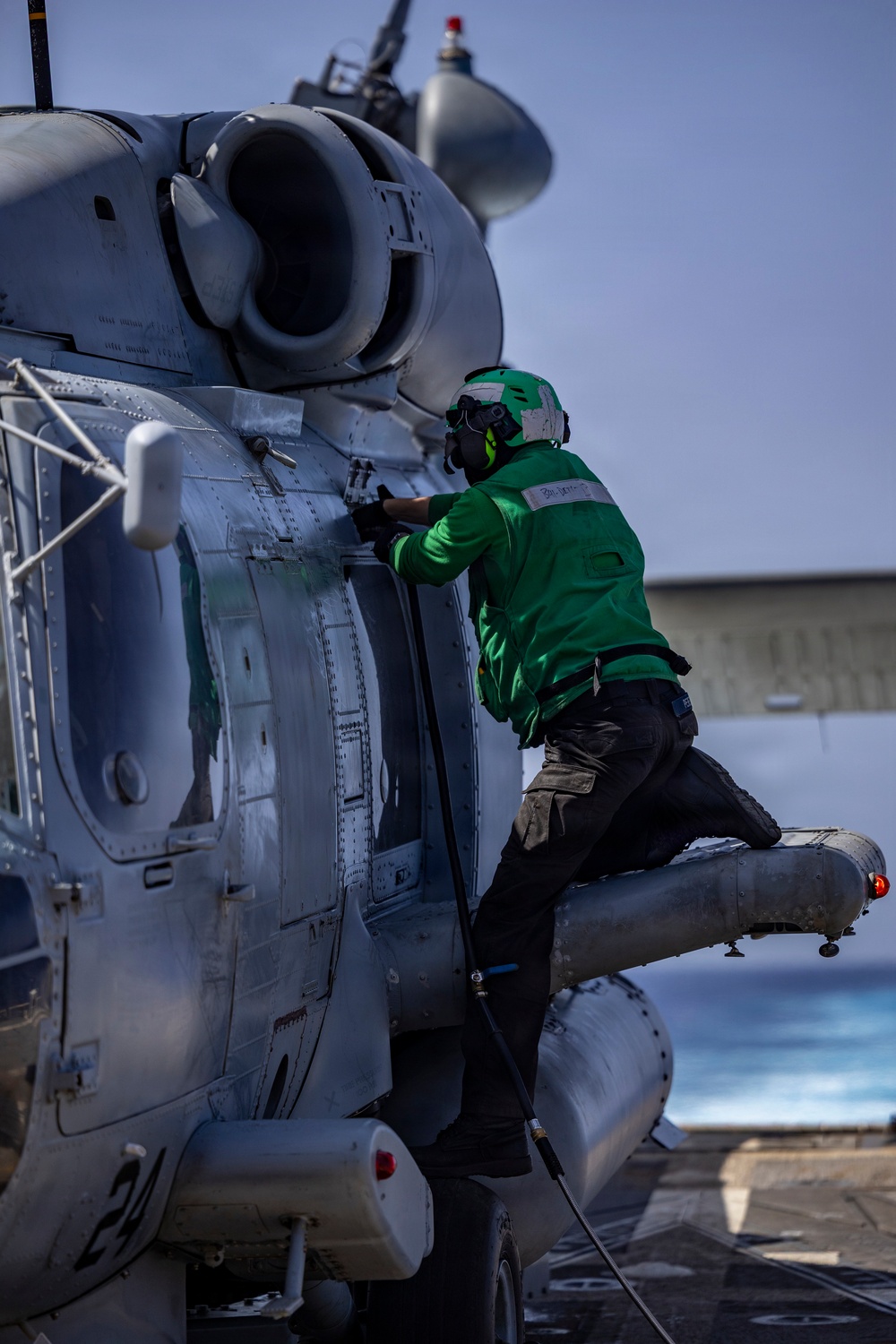 A naval aviation technician in a green jersey and helmet is performing maintenance or inspection work on a military helicopter, likely an MH-60 Seahawk, aboard what appears to be a naval vessel. The technician is climbing on the helicopter's sponson/float while working on the fuselage near the engine compartment. The setting appears to be a ship's flight deck with ocean visible in the background.