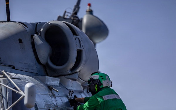 A naval aviation technician in a green jersey and helmet is performing maintenance or inspection work on a military helicopter, likely an MH-60 Seahawk, aboard what appears to be a naval vessel. The technician is climbing on the helicopter's sponson/float while working on the fuselage near the engine compartment. The setting appears to be a ship's flight deck with ocean visible in the background.
