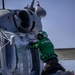 A naval aviation technician in a green jersey and helmet is performing maintenance or inspection work on a military helicopter, likely an MH-60 Seahawk, aboard what appears to be a naval vessel. The technician is climbing on the helicopter's sponson/float while working on the fuselage near the engine compartment. The setting appears to be a ship's flight deck with ocean visible in the background.