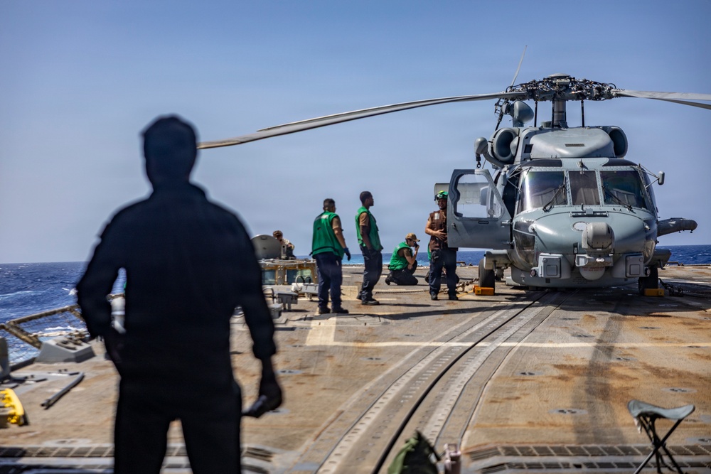 Military personnel are conducting maintenance or inspection operations on a naval helicopter aboard a ship's flight deck at sea. Several crew members wearing green vests are working around what appears to be an MH-60 Seahawk helicopter secured to the deck. The scene takes place on a clear day with open ocean visible in the background.