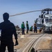 Military personnel are conducting maintenance or inspection operations on a naval helicopter aboard a ship's flight deck at sea. Several crew members wearing green vests are working around what appears to be an MH-60 Seahawk helicopter secured to the deck. The scene takes place on a clear day with open ocean visible in the background.