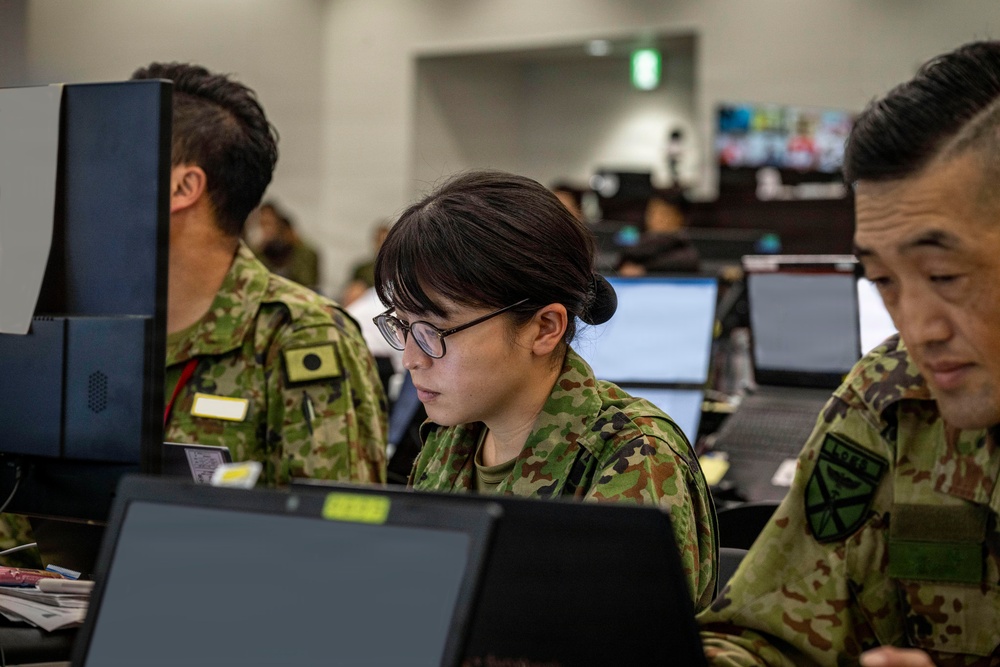 Three military personnel in Japanese Ground Self-Defense Force (JGSDF) camouflage uniforms are working at computer workstations in what appears to be a command or operations center. The central figure is a female soldier wearing glasses, focused on a laptop screen. The setting suggests a military exercise or operational planning environment.