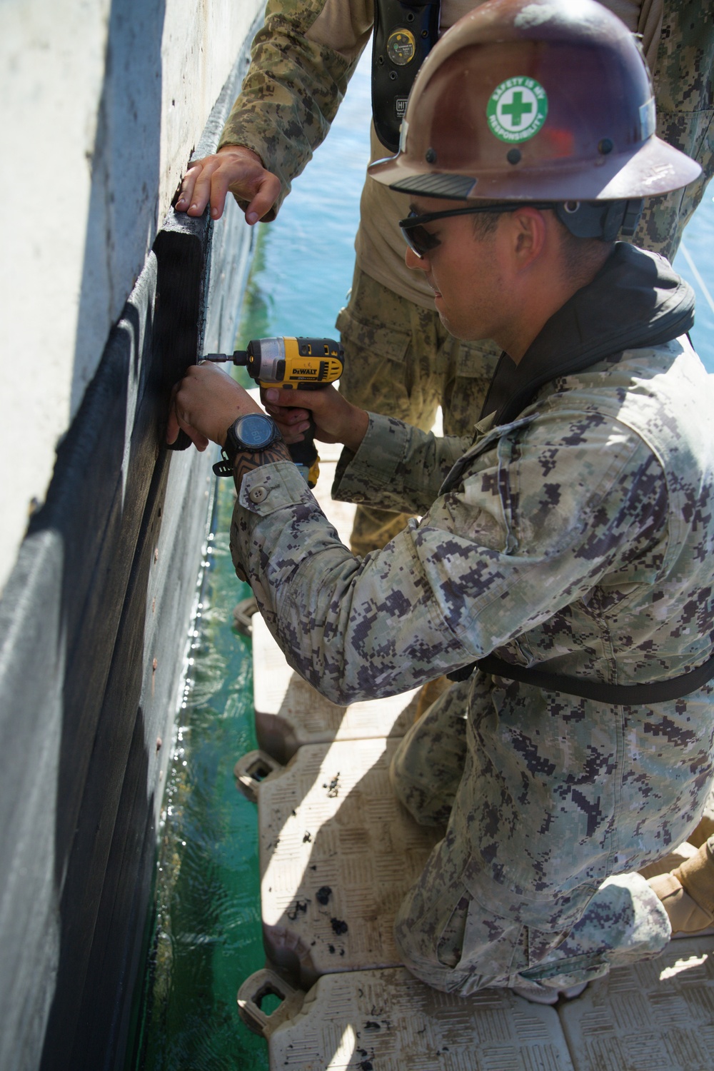 A military service member wearing Navy Working Uniform (NWU) digital camouflage and a hard hat with a 'Safety Is My Responsibility' sticker is using a DeWalt power drill to perform maintenance or repair work on the hull of a vessel at the waterline. A second person in military camouflage is visible above, assisting by holding the work surface. The work is being conducted from a small platform or pontoon adjacent to the vessel over open water.