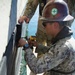 A military service member wearing Navy Working Uniform (NWU) digital camouflage and a hard hat with a 'Safety Is My Responsibility' sticker is using a DeWalt power drill to perform maintenance or repair work on the hull of a vessel at the waterline. A second person in military camouflage is visible above, assisting by holding the work surface. The work is being conducted from a small platform or pontoon adjacent to the vessel over open water.