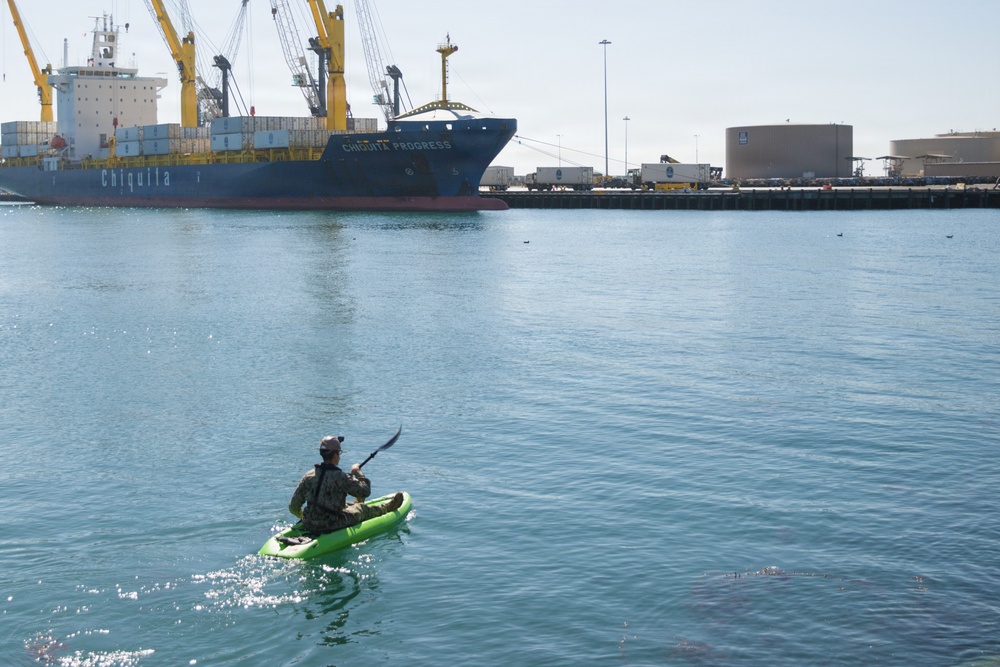 A person in camouflage clothing is kayaking on a green inflatable kayak in a commercial harbor. In the background, a large Chiquita cargo ship named 'Chiquita Progress' is docked at a port facility with yellow cranes and industrial infrastructure visible. The contrast between the small recreational kayaker and the massive commercial vessel creates a striking visual juxtaposition.