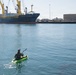 A person in camouflage clothing is kayaking on a green inflatable kayak in a commercial harbor. In the background, a large Chiquita cargo ship named 'Chiquita Progress' is docked at a port facility with yellow cranes and industrial infrastructure visible. The contrast between the small recreational kayaker and the massive commercial vessel creates a striking visual juxtaposition.
