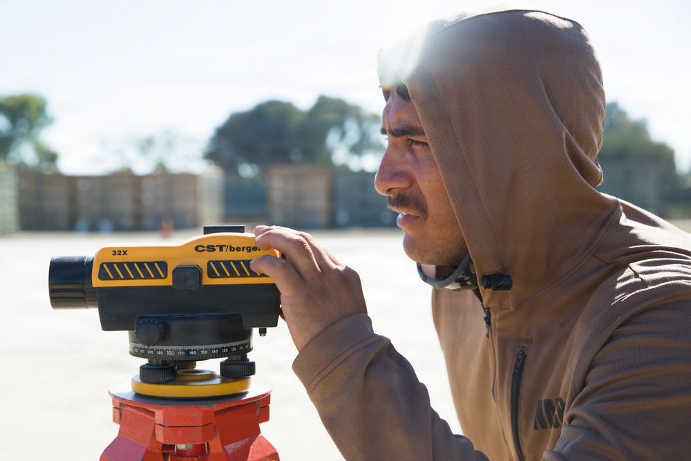A man wearing a tan/brown hooded sweatshirt is operating a CST/Berger 32X automatic optical level surveying instrument mounted on a red tripod. He appears to be looking through or adjusting the instrument in an open outdoor area. The background shows a paved surface and blurred structures or barriers.