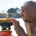 A man wearing a tan/brown hooded sweatshirt is operating a CST/Berger 32X automatic optical level surveying instrument mounted on a red tripod. He appears to be looking through or adjusting the instrument in an open outdoor area. The background shows a paved surface and blurred structures or barriers.