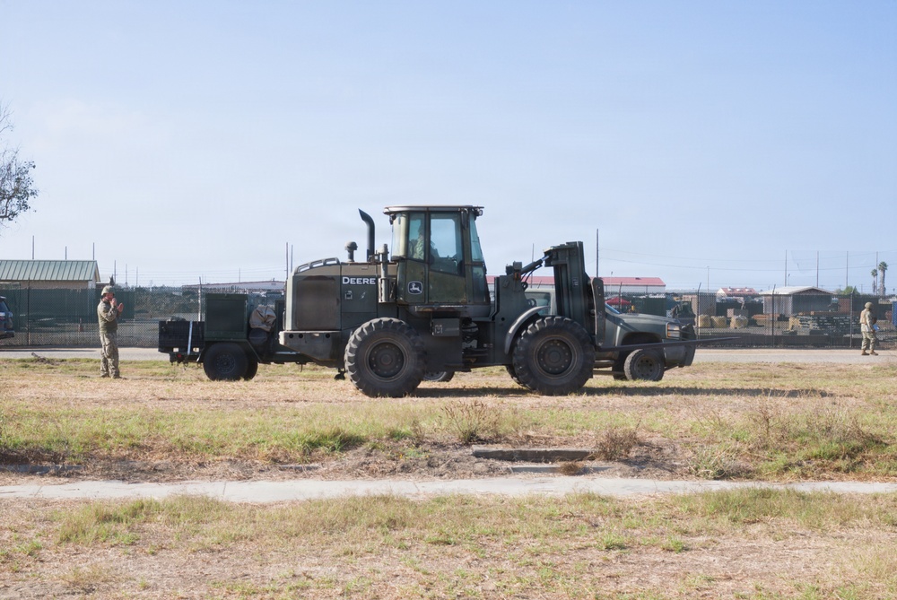 A military-painted John Deere wheel loader/forklift is positioned on a grassy field within what appears to be a military installation or base. Two personnel in military uniforms are visible, one on the left appearing to observe or communicate, and one on the far right. The background shows fenced compound areas with various structures and equipment storage.