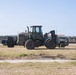 A military-painted John Deere wheel loader/forklift is positioned on a grassy field within what appears to be a military installation or base. Two personnel in military uniforms are visible, one on the left appearing to observe or communicate, and one on the far right. The background shows fenced compound areas with various structures and equipment storage.