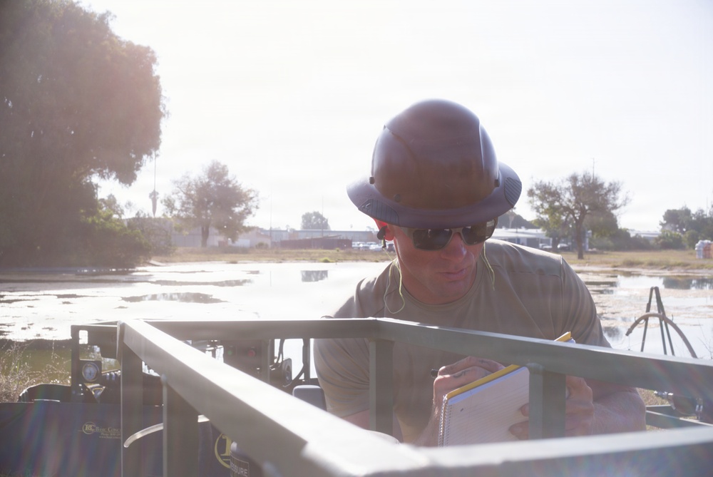 A worker wearing a full-brim hard hat and sunglasses is writing in a notepad while positioned near or inside a piece of equipment or vehicle. The setting appears to be an outdoor work site with standing water visible in the background, suggesting possible flood or construction conditions. Bright backlighting creates lens flare and reduces overall image contrast.