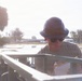 A worker wearing a full-brim hard hat and sunglasses is writing in a notepad while positioned near or inside a piece of equipment or vehicle. The setting appears to be an outdoor work site with standing water visible in the background, suggesting possible flood or construction conditions. Bright backlighting creates lens flare and reduces overall image contrast.