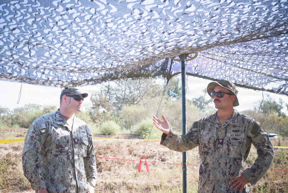 Two U.S. Navy Seabees personnel are standing under a camouflage netting canopy in an outdoor field environment. The individual on the right, whose name tape reads 'LEE' and whose uniform bears 'U.S. NAVY' markings, appears to be briefing or explaining something to the individual on the left, whose name tape reads 'McCOURIE'. Both are wearing standard U.S. Navy Working Uniform (NWU) with sunglasses.