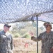Two U.S. Navy Seabees personnel are standing under a camouflage netting canopy in an outdoor field environment. The individual on the right, whose name tape reads 'LEE' and whose uniform bears 'U.S. NAVY' markings, appears to be briefing or explaining something to the individual on the left, whose name tape reads 'McCOURIE'. Both are wearing standard U.S. Navy Working Uniform (NWU) with sunglasses.